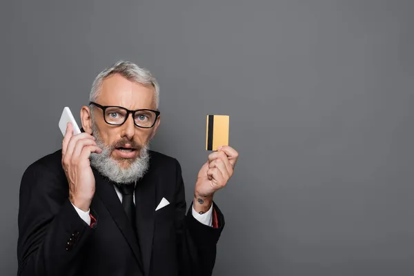 Überraschter Geschäftsmann mittleren Alters mit Kreditkarte, während er auf dem Smartphone isoliert auf grau spricht — Stockfoto