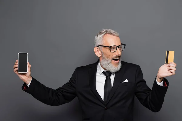 Sorprendido hombre de negocios de mediana edad con tarjeta de crédito y teléfono inteligente con pantalla en blanco en gris - foto de stock