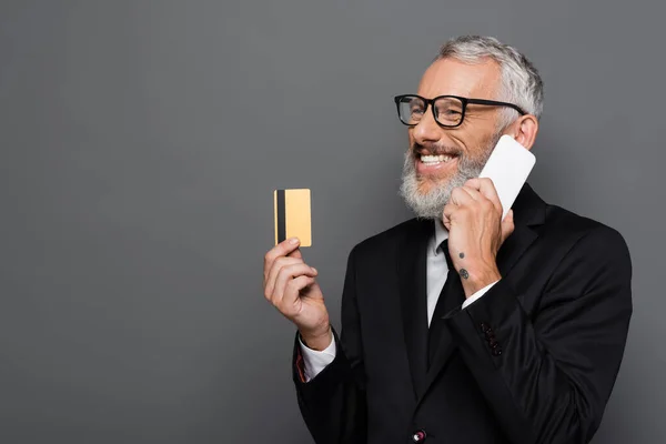 Glücklicher Geschäftsmann mittleren Alters mit Kreditkarte, während er auf dem Smartphone isoliert auf grau spricht — Stockfoto