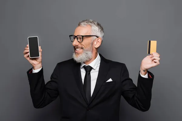 Feliz hombre de negocios de mediana edad con tarjeta de crédito y teléfono inteligente con pantalla en blanco en gris - foto de stock