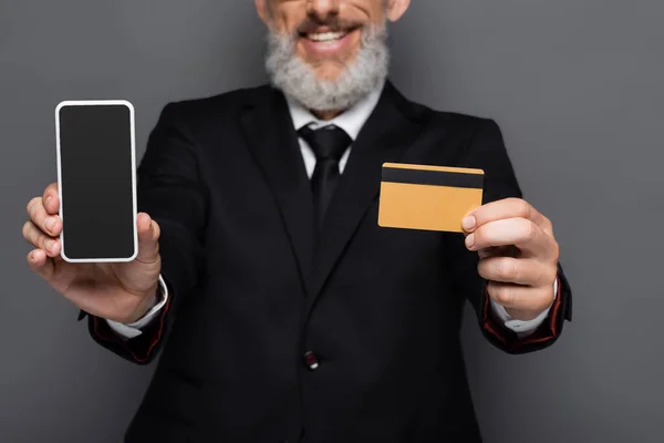 Обрезанный вид счастливого бизнесмена средних лет, держащего кредитную карту и смартфон с пустым экраном, изолированным на сером — стоковое фото