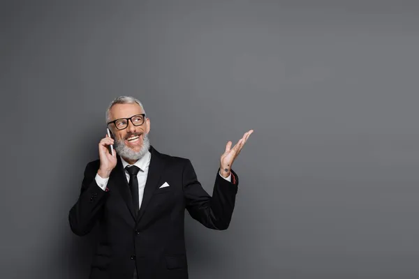 Alegre hombre de negocios de mediana edad hablando por teléfono celular y haciendo gestos en gris - foto de stock