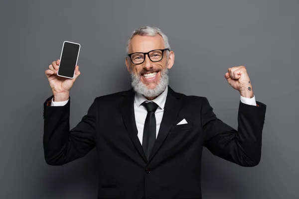 Cheerful middle aged businessman holding cellphone with blank screen on grey — Stock Photo