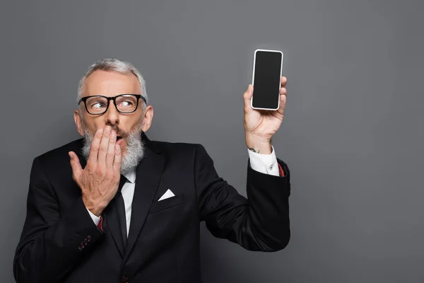 Middle aged businessman holding cellphone with blank screen and covering mouth on grey — Stock Photo