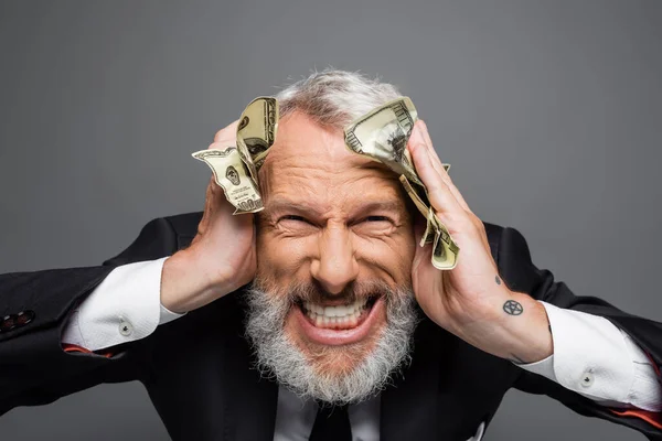 Loco empresario de mediana edad sosteniendo billetes de dólar y sonriendo aislado en gris - foto de stock