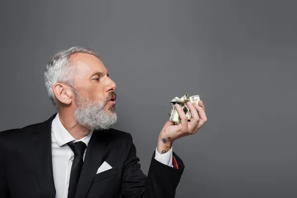 Вид збоку бородатого середнього віку бізнесмена, який дме на збиті доларові банкноти ізольовані на сірому — стокове фото