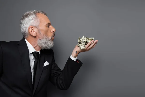 Side view of tattooed middle aged businessman blowing on crumpled dollar banknotes on grey — Stock Photo