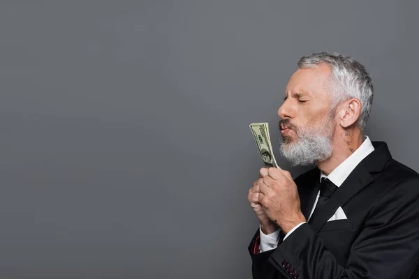 Татуйований бізнесмен середнього віку цілує доларові банкноти ізольовані на сірому — стокове фото