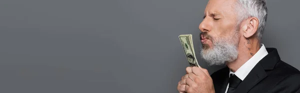 Hombre de negocios tatuado de mediana edad besando billetes de dólar aislados en gris, pancarta - foto de stock