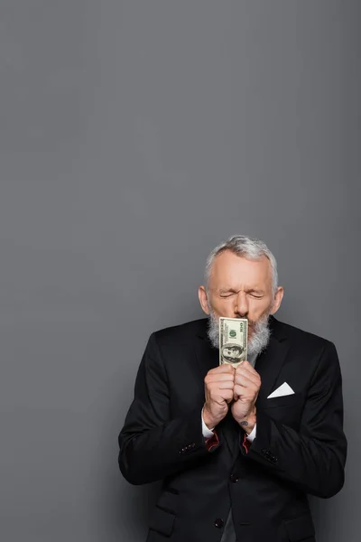 Татуйований бізнесмен середнього віку з закритими очима цілує доларові банкноти на сірому — стокове фото