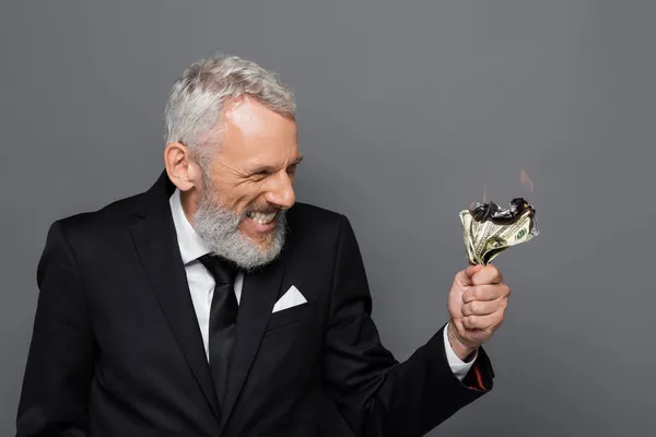 Happy middle aged businessman holding burning dollar banknotes isolated on grey — Stock Photo