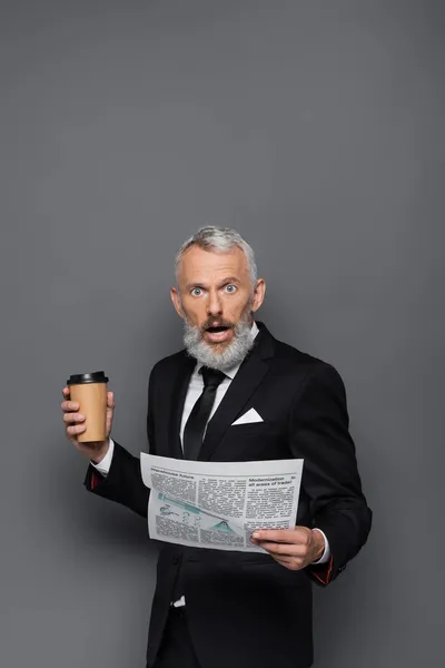 Homem de negócios de meia idade chocado segurando copo de papel e jornal em cinza — Fotografia de Stock