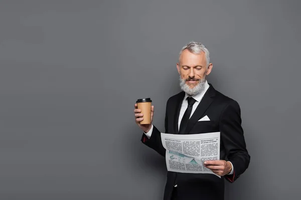 Sonriente hombre de negocios de mediana edad sosteniendo la taza de papel y leyendo el periódico en gris - foto de stock