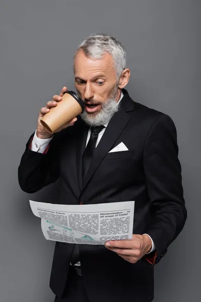Шокированный бизнесмен средних лет, держащий бумажный стаканчик и читающий газету на сером — стоковое фото