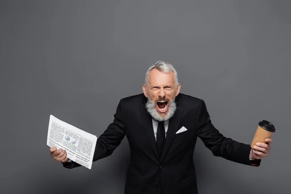 Angry middle aged businessman holding paper cup and newspaper while screaming on grey — Stock Photo