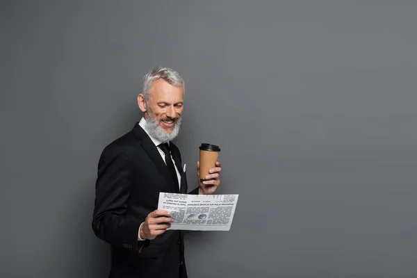 Alegre hombre de negocios de mediana edad sosteniendo la taza de papel y leyendo el periódico en gris - foto de stock