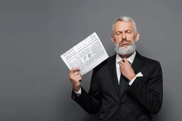 Middle aged businessman adjusting tie and holding newspaper on grey — Stock Photo