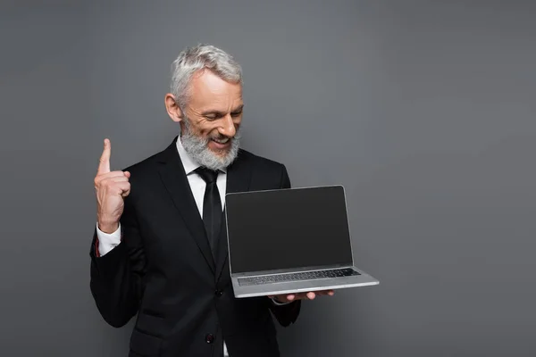 Happy middle aged businessman in suit holding laptop with blank screen and pointing up isolated on grey — Stock Photo