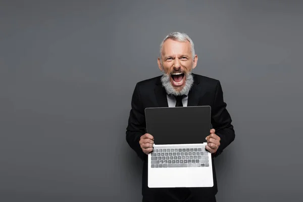 Amazed middle aged businessman in suit holding laptop with blank screen on grey — Stock Photo