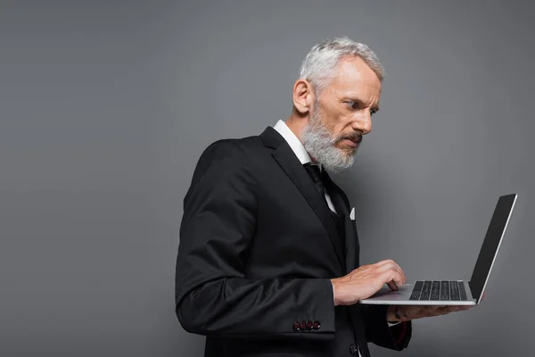 Homme d'affaires d'âge moyen en costume à l'aide d'un ordinateur portable avec écran blanc isolé sur gris — Photo de stock