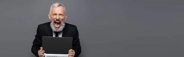 Здивований середнього віку бізнесмен у костюмі тримає ноутбук з порожнім екраном на сірому, банер — стокове фото