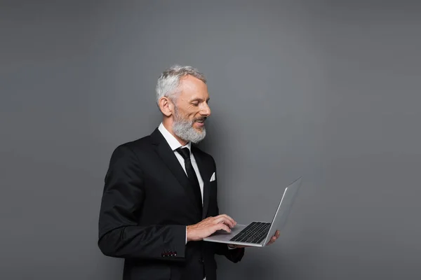 Hombre de negocios de mediana edad feliz en traje usando el ordenador portátil en gris - foto de stock