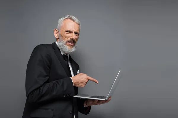 Hombre de negocios de mediana edad en traje apuntando con el dedo a la computadora portátil aislado en gris - foto de stock