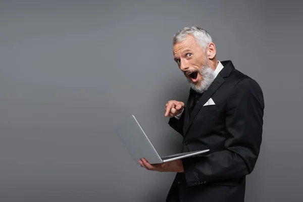 Sorprendido hombre de negocios de mediana edad en traje apuntando con el dedo a la computadora portátil en gris - foto de stock