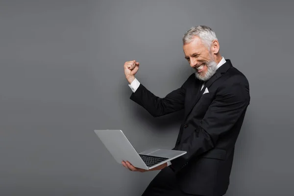 Excited middle aged businessman in suit holding laptop on grey — Stock Photo