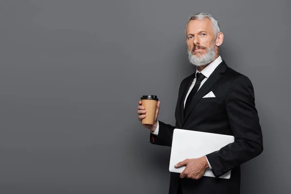 Hombre de negocios de mediana edad en traje que sostiene el ordenador portátil y la taza de papel en gris - foto de stock