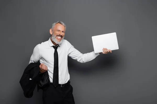 Happy middle aged businessman holding laptop and blazer on grey — Stock Photo