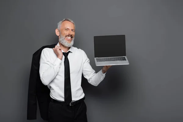 Happy middle aged businessman in suit holding laptop with blank screen and blazer on grey — Stock Photo