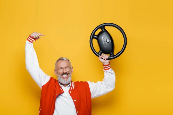 Happy middle aged man in bomber jacket pointing at steering wheel on yellow — Stock Photo