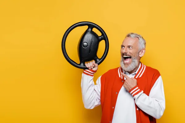 Amazed middle aged man in bomber jacket pointing at steering wheel on yellow — Stock Photo