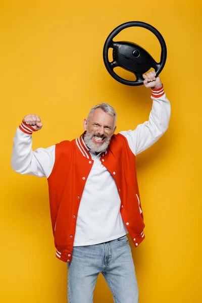 Happy middle aged man in bomber jacket holding steering wheel above head on yellow — Stock Photo