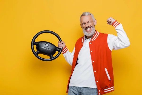 Excited middle aged man in bomber jacket holding steering wheel on yellow — Stock Photo