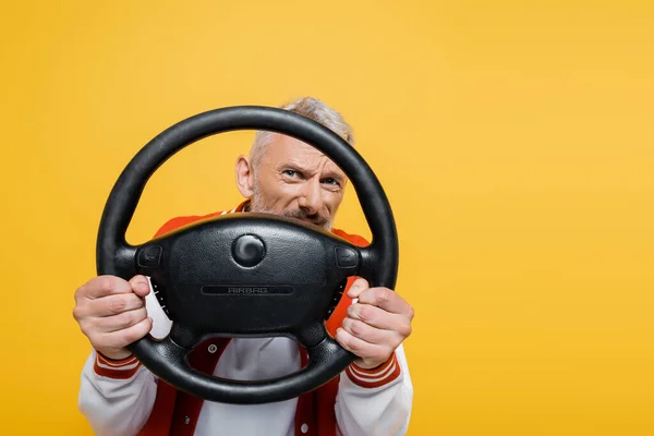 Playful middle aged man in bomber jacket holding steering wheel isolated on yellow — Stock Photo