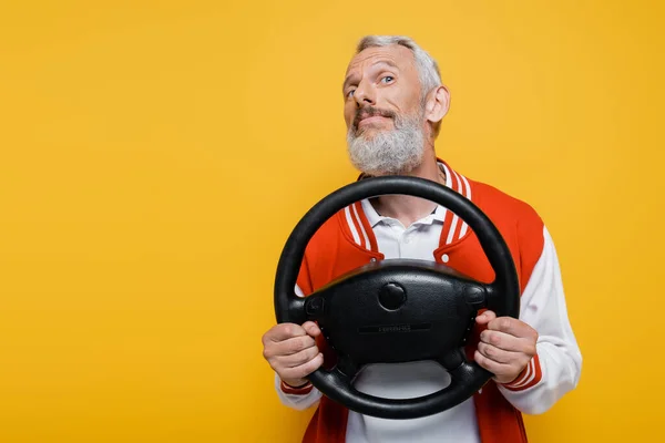 Soddisfatto uomo di mezza età in giacca bomber che tiene il volante mentre imita la guida isolata sul giallo — Foto stock