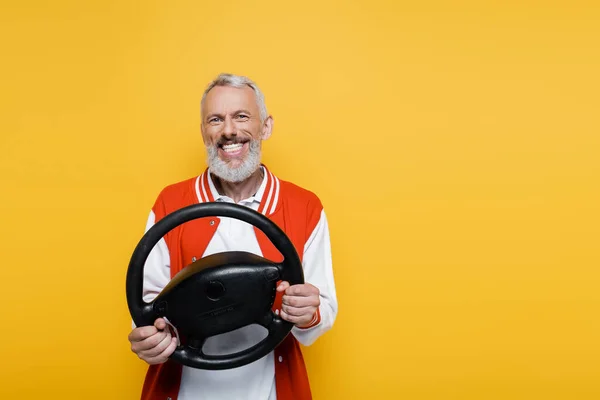 Joyful middle aged man in bomber jacket holding steering wheel while imitating driving isolated on yellow — Stock Photo