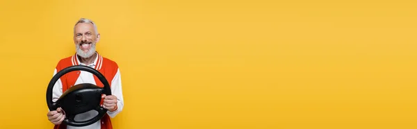 Felice uomo di mezza età in giacca bomber che tiene il volante mentre imita la guida isolata sul giallo, banner — Foto stock