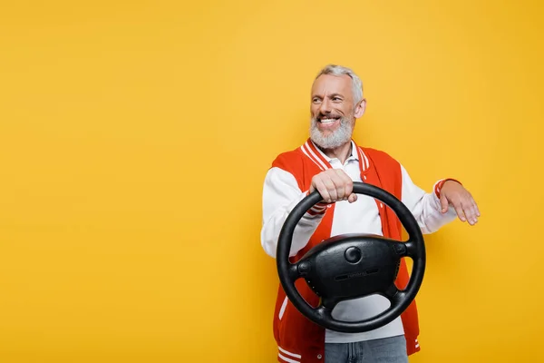 Glücklicher Mann mittleren Alters in Bomberjacke hält das Lenkrad, während er isoliert auf gelb gestikuliert — Stockfoto