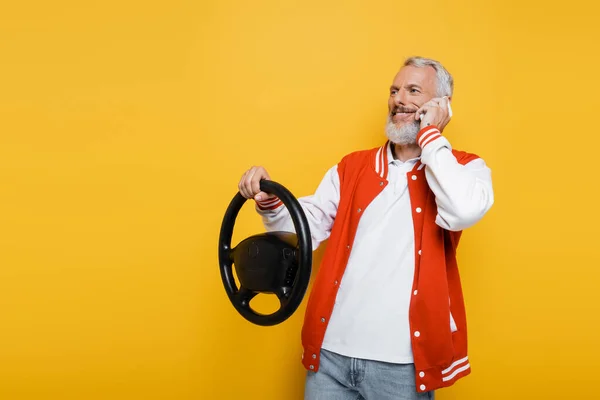 Felice uomo di mezza età che tiene il volante mentre imita la guida e parla su smartphone isolato sul giallo — Foto stock