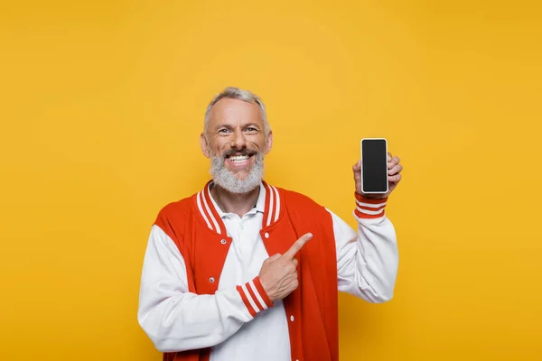 Hombre de mediana edad feliz apuntando al teléfono inteligente con pantalla en blanco aislado en amarillo - foto de stock