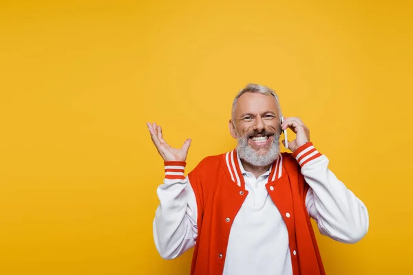 Glücklicher Mann mittleren Alters spricht auf Smartphone und gestikuliert isoliert auf gelb — Stockfoto