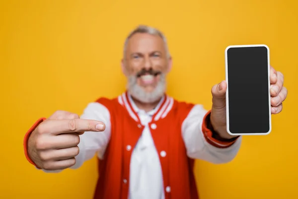 Размытый и счастливый мужчина средних лет указывает на смартфон с пустым экраном, изолированным на желтом — стоковое фото