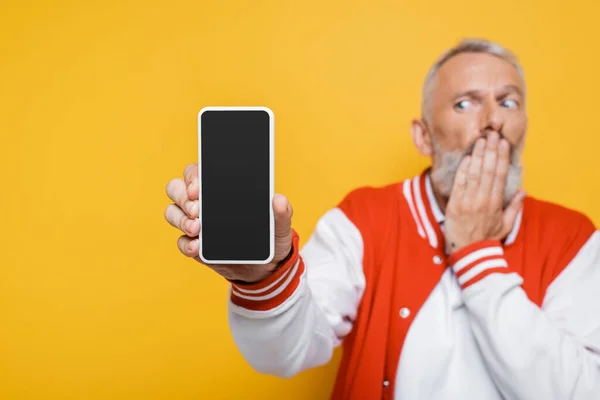 Blurred middle aged man holding smartphone with blank screen and covering mouth isolated on yellow — Stock Photo