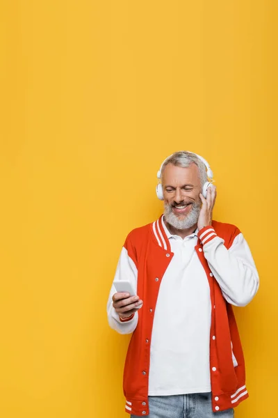 Positive middle aged man in wireless headphones holding cellphone and listening music isolated on yellow — Stock Photo