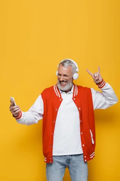 Hombre positivo de mediana edad en auriculares tomando selfie mientras muestra el signo de roca en amarillo - foto de stock