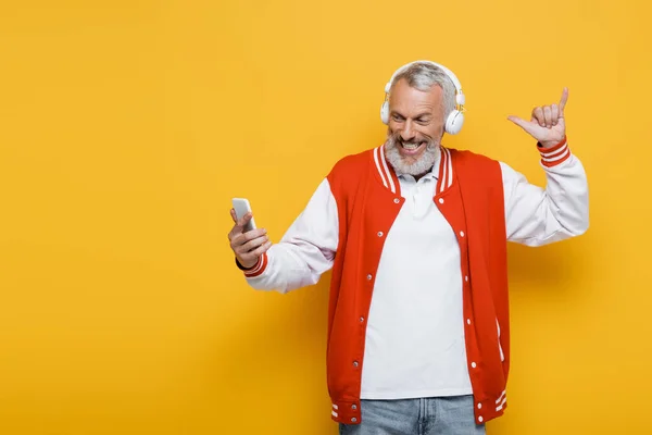 Positive middle aged man in headphones taking selfie while showing hang loose gesture on yellow — Stock Photo