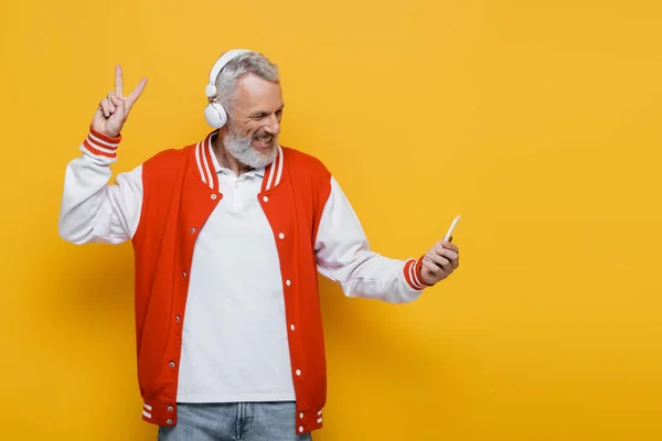 Feliz homem de meia idade em fones de ouvido tomando selfie enquanto mostra o sinal da peça no amarelo — Fotografia de Stock